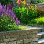 A stone wall in the garden with flowers coming out of the top.